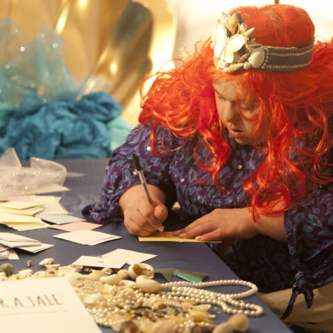 A woman writes on cards, while surrounded by props and jewellery.