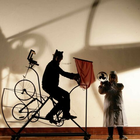 A person riding a bicycle type machine, a person holding an electric fan. Shadows cast on the wall.