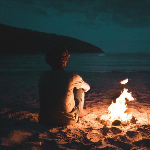 A man sits besides a small fire on beach at night