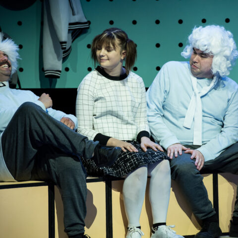 Three people sit on stage, laughing and smiling. The two men are wearing white wigs. A girl in pigtails sits in the middle.
