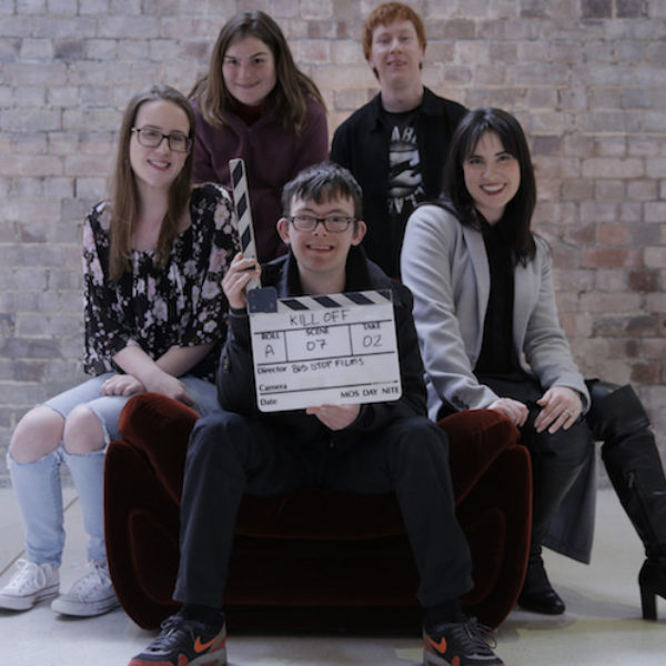 Young man holding clapper board surrounded by three women and another young man.