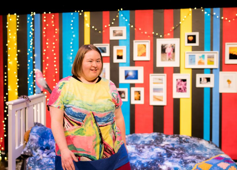 Artist is in the centre of her set designed bedroom, artist is wearing a bright top with rainbow colours. Behind artist is a bed frame with doona cover of stars. Behind the bed is a wall painted in red, black, blue and yellow strips. On the wall are a series of photographs . They are hung on the wall in different sized frames. 