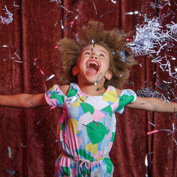 Young girl in standing in front of red curtain throwing shredded glittery paper in the air, with her mouth open and arms outstretched