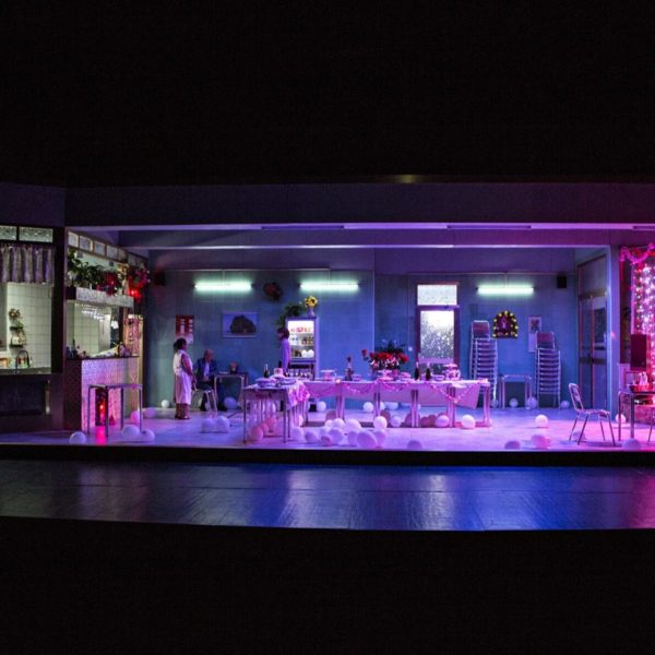 Stage with stark restaurant set up for an event, 2 people in the restaurant area, 1 performer to the left in the kitchen, 1 to the right on a small stage singing into a microphone