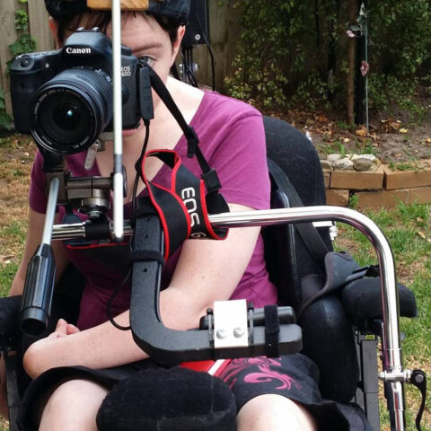 Woman in wheelchair in pink shirt looking through a camera lens