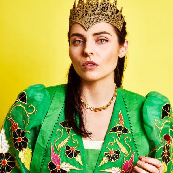 Ange Lavoipierre wears a bold crown and heavily embroidered green dress in front of a vivid yellow background.