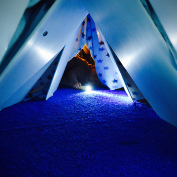 A blue blanket fort on blue carpet. In the middle of the image, a child is lying at the fort entrance shining a torch at the camera. Their face is obscured. There is warm yellow light glowing behind them.