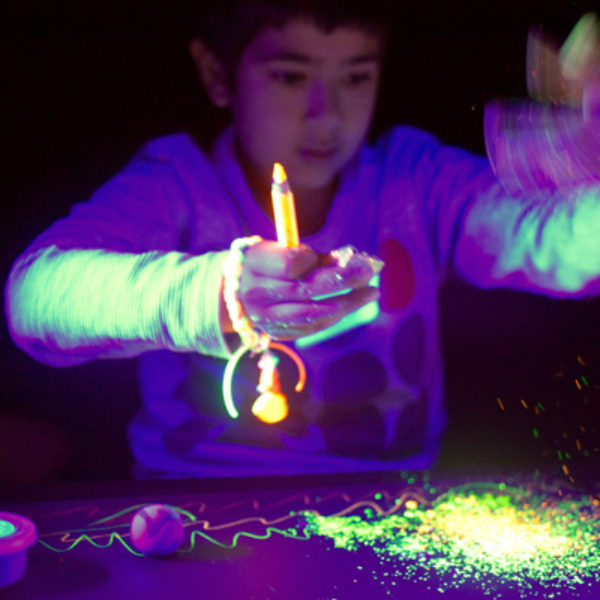 A child behind a table, looking at a pile of glowing particles, holding their hands in front of them. Their left hand is a motion blur as they are shaking it. They hold a yellow pencil and some other toys in their right hand.