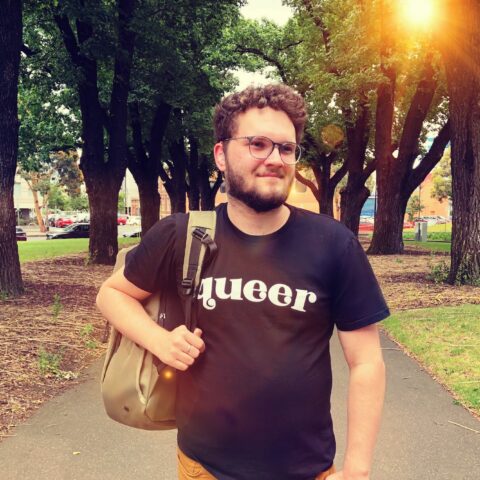 Sam Martin, a young caucasian man wearing black tee shirt with “queer” in white letters and a clear hearing aid, walks through a park