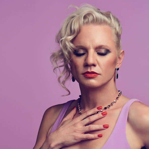 A stunning trans woman in light purple holds her hand to her chest while looking down showcasing her dramatic eye make up