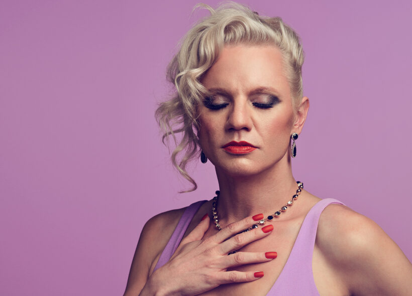A stunning trans woman in light purple holds her hand to her chest while looking down showcasing her dramatic eye make up