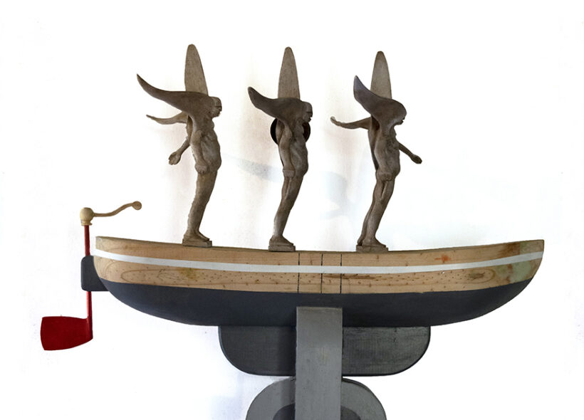Three light brown winged male figures stand on a wooden boat like shape with the bottom painted grey. The rudder is painted red. 