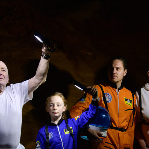 A family — man in his sixties carrying a space helmet, an 11 year old girl wearing a blue space suit, a man in his 30s in an orange space suit carrying a space helmet, and a woman in her 50s stand against an alien looking, rock wall. They hold lights aloft in the darkness.