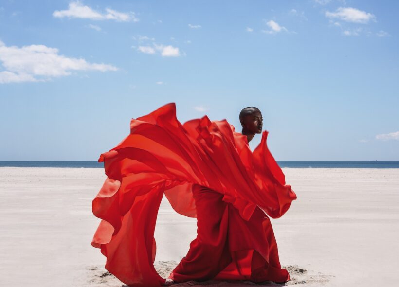 A black woman in an elegant red dress as standing on the beach. Her dress is flying around her. 