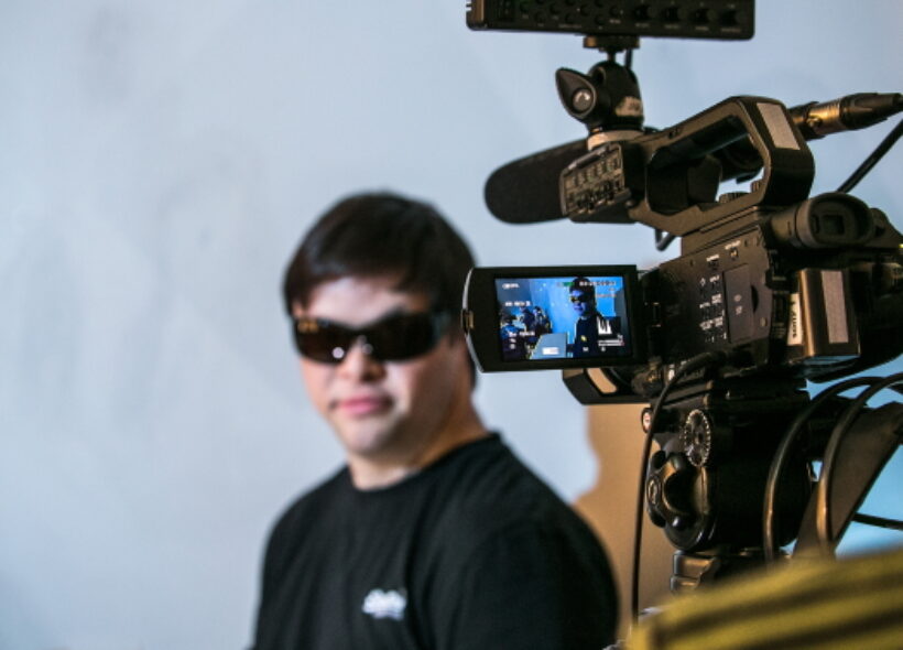 A man in black clothes and sunglasses is filmed by a camera