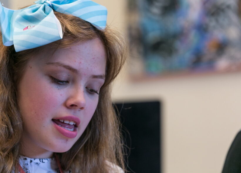 A photo of a girl with a blue bow in her hair. Her eyes are cast downwards as she sings.