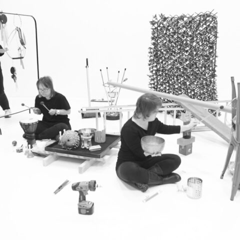 Three musicians are creating sound within an installation of various objects and hanging instruments.