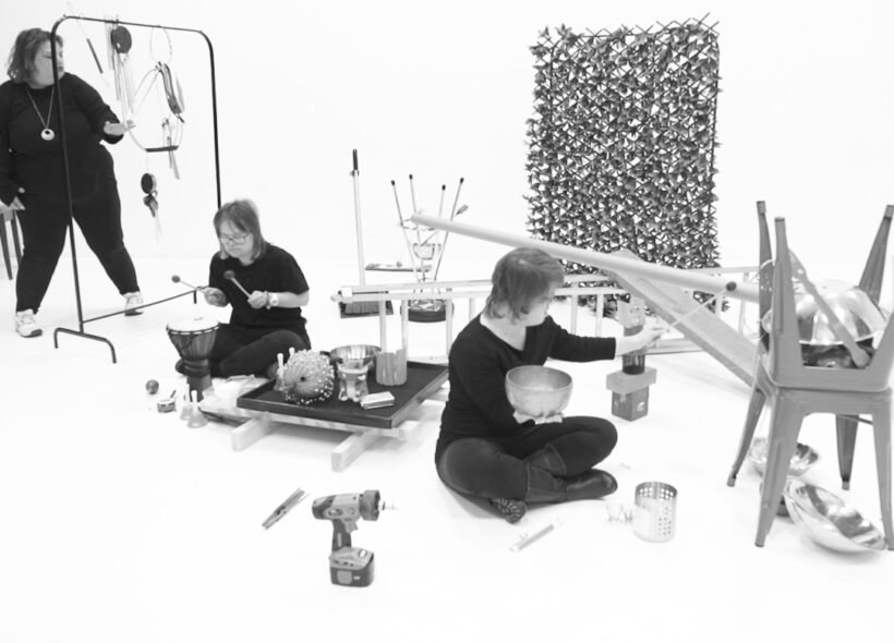 Three musicians are creating sound within an installation of various objects and hanging instruments.