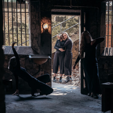There are four dancers dressed in black, pictured in the historic underground J Ward Asylum kitchen surrounded by bluestone floors and walls. The windows are barred, with an old gas light emitting a soft glow that pierces through the cold, dark interior. Two of the dancers in the foreground move in tableau through the darkness, and two dancers can be seen embracing through the open doorway in the light of day.