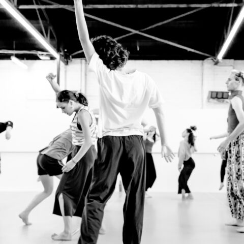 Black & white image of dancers moving variously in a white walled studio