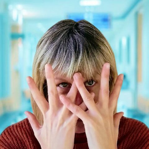 Woman's face with two hands stretched over eyes, in blurry blue hospital corridor