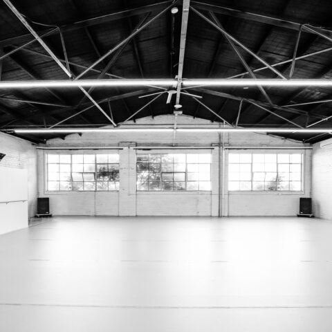 Image shows a dance studio in a warehouse space, with thin light beams hanging from a black, sloped ceiling, wide windows and white brick walls