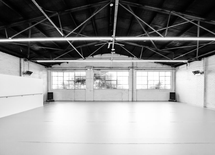 Image shows a dance studio in a warehouse space, with thin light beams hanging from a black, sloped ceiling, wide windows and white brick walls