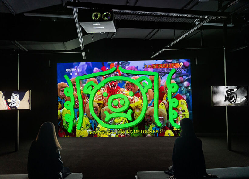 Two people sitting on a bench looking a large screen. The screen has colourful patterns with a neon green painting on top depicting a pig.