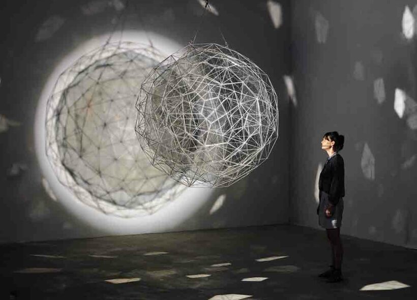 A person standing in front of the artwork 'Stardust particle' by Olafur Eliasson. The artwork is a large spherical net hanging from the ceiling and refracting pale white light in a dark room.
