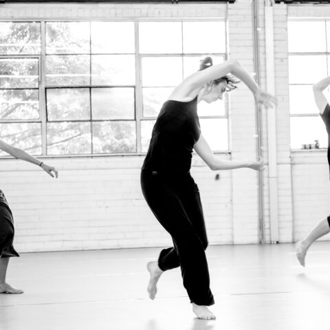 3 dancers move dynamically in front of a large window in a greyscale image.