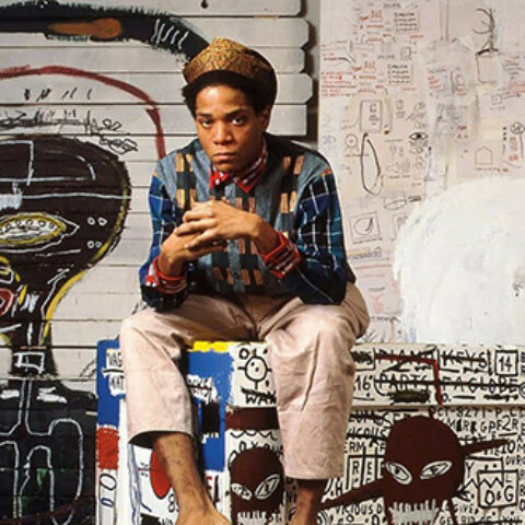 Photo of young Jean-Michel Basquiat sitting on a bench with his hands clasped together. The background and the bench are covered in his paintings.