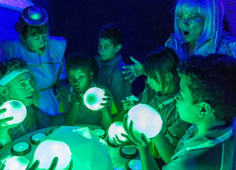 Four young children in school uniform surround a table along with three adults dressed in silver costume. One adult and two children are holding a lit orb. They are all lit in green and purple lighting. 