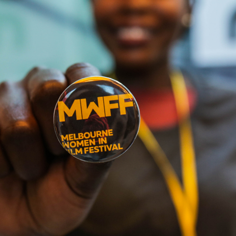 A smiling woman holding out a promotional button in the foreground of the image that has the logo for the Melbourne Women in Film Festival on it.