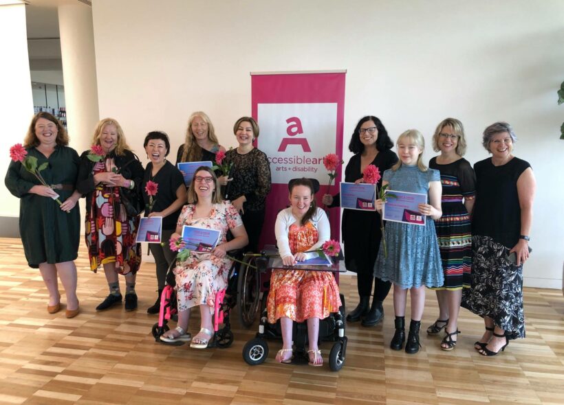 Eleven women in front of a pink Accessible Arts banner. They are smiling and holding pink flowers and certificates.