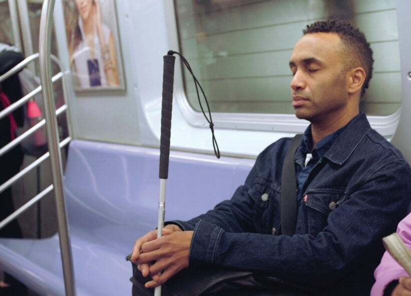 A picture of film maker Rodney Evans sitting on a blue seat on a train. The image is cut off at the waist. Rodney holds his cane in front of him with two hands, his eyes are closed. Rodney has short brown hair, he is wearing a dark blue shirt and a slightly darker denim jacket. Behind him there is a window, advertising posters and other people riding the train.