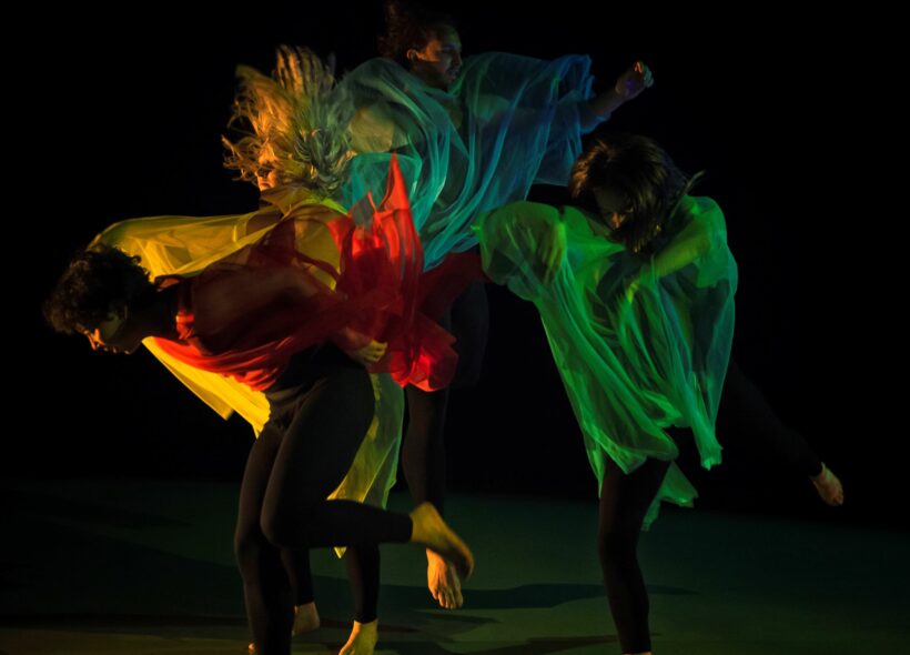 Four dancers dancing together against a black backdrop, facing different directions. They have been captured in motion and have their eyes closed. They are wrapped in swirling pieces of sheer fabric, and are lit from the sides with yellow, red, teal, blue and green light. 