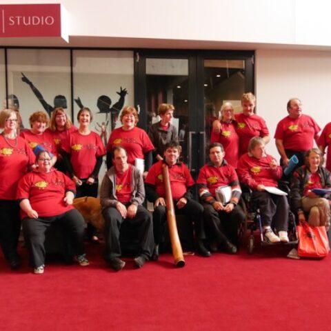 choir members wearing bright red T shirts in two rows. The front row sitting and the back row standing. A man in the centre holds a didgeridoo, another at the end holds a guitar