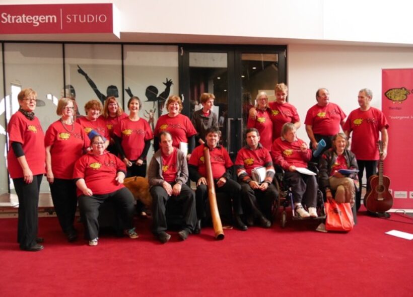 choir members wearing bright red T shirts in two rows. The front row sitting and the back row standing. A man in the centre holds a didgeridoo, another at the end holds a guitar