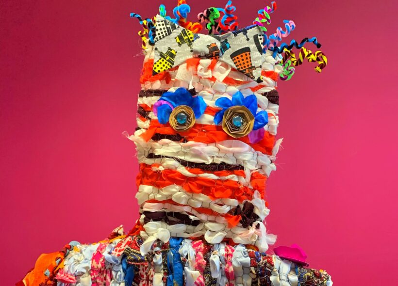 Fabric sculpture of stylised head and shoulders. Strips of very bright coloured cloth, button eyes with bright blue lashes give a wild and zany effect.