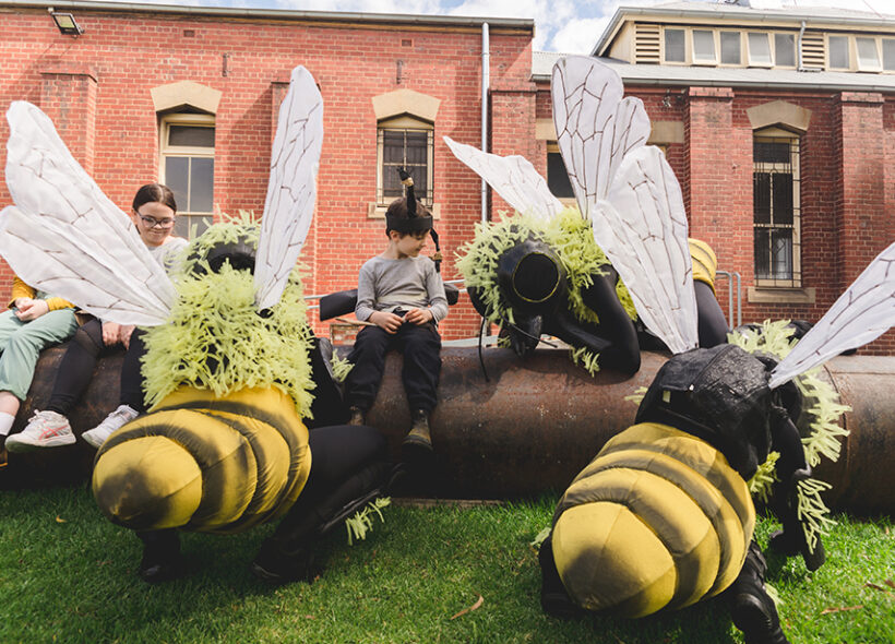 A Bees production photo. Polyglot artists in intricate Bee costumes are perched on and around a large metal pipe on green lawn. Children wearing handmade paper antennae and wings sit on the pipe amongst them. There is a heritage red brick building in the background.