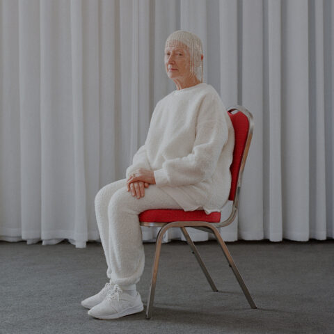 An older person dressed in a white tracksuit, white sneakers, and a headpiece made of strands of diamentes sits on a red chair, which is placed on a dark grey carpet. The chair is placed in front of long light grey drapes. 