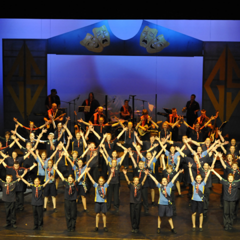 A large group of scouts perform a choreographed routine on stage, with hands raised in a V-shape. The background features theatrical masks and musical instruments.