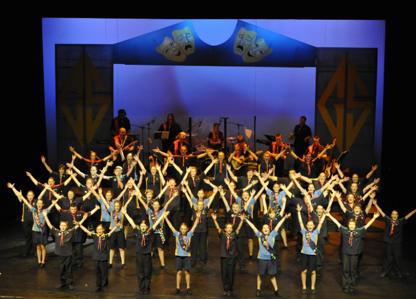 A large group of scouts perform a choreographed routine on stage, with hands raised in a V-shape. The background features theatrical masks and musical instruments.
