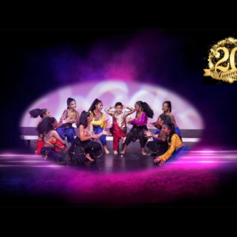 A group of dancers performs energetically on stage, dressed in vibrant costumes. They are arranged in a dynamic formation, striking expressive poses. The background features colourful, ambient lighting with purple and pink hues, creating a lively atmosphere.