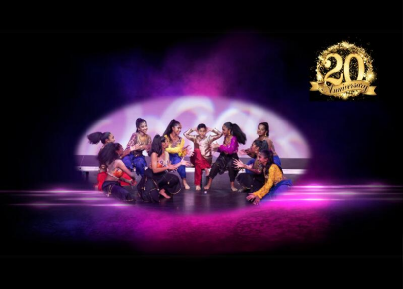 A group of dancers performs energetically on stage, dressed in vibrant costumes. They are arranged in a dynamic formation, striking expressive poses. The background features colourful, ambient lighting with purple and pink hues, creating a lively atmosphere.