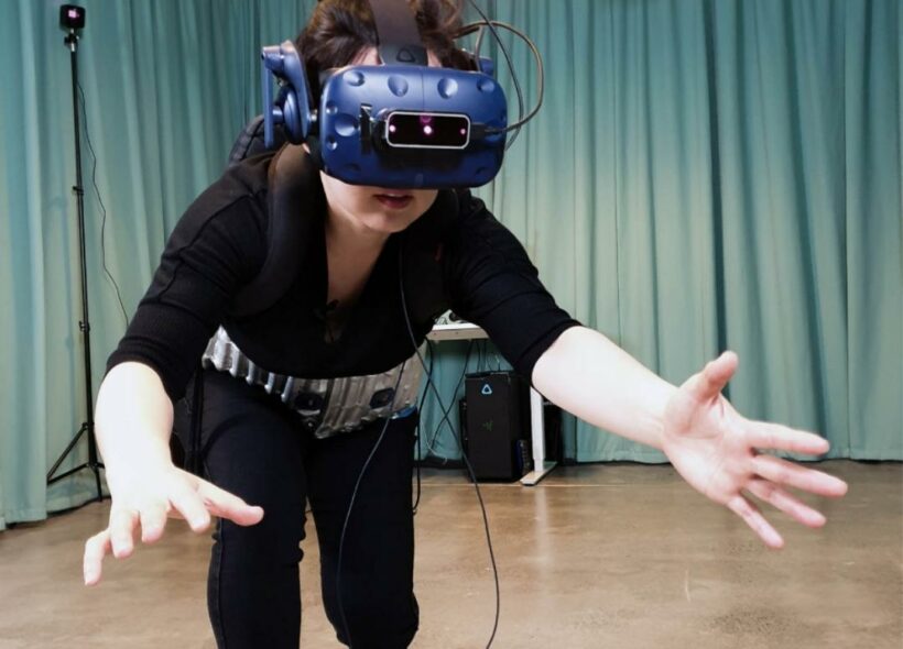  A person wearing headgear and a large custom-built belt is engaging in a virtual reality game. They are bending over with both arms stretched forwards. Interdisciplinary artwork credit ‘Breakout My Pelvic Sorcery’ by Eugenie Lee.