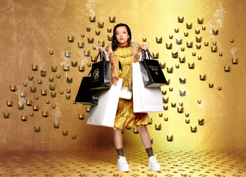 Woman in yellow dress, against gold background, holding many designer label shopping bags