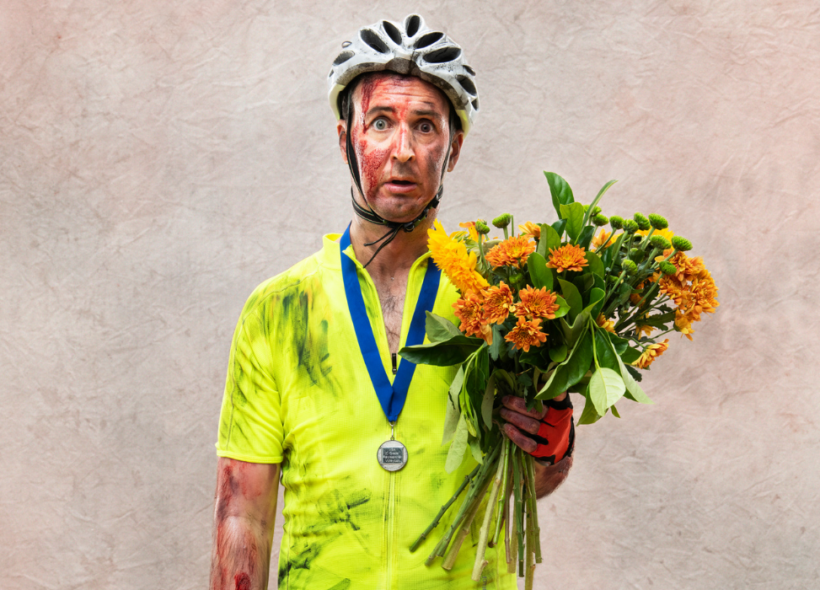 A man stands dressed in a helmet a full cycling clothing, he is holding a bunch of flowers and looks bruised and battered like he has been in a crash.