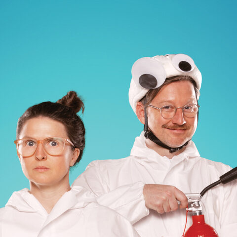 Two people wearing white onsies, shown from the shoulders up, stand, one slightly behind the other. One has their long hair up in a high bun and glasses - they are looking quizzical. One has glasses and a moustache and is smiling widely. They are wearing a white hat with large googly eyes on it and are setting off a red fire extinguisher. The background is teal.