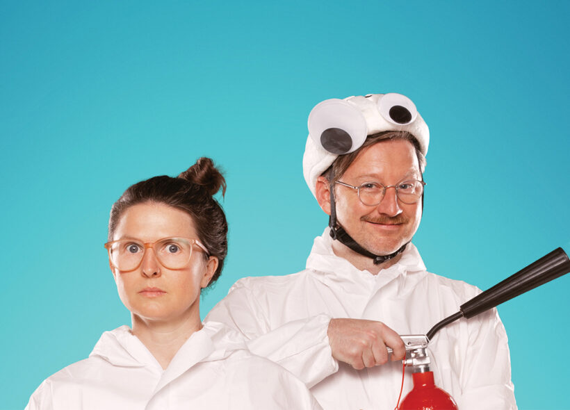 Two people wearing white onsies, shown from the shoulders up, stand, one slightly behind the other. One has their long hair up in a high bun and glasses - they are looking quizzical. One has glasses and a moustache and is smiling widely. They are wearing a white hat with large googly eyes on it and are setting off a red fire extinguisher. The background is teal.
